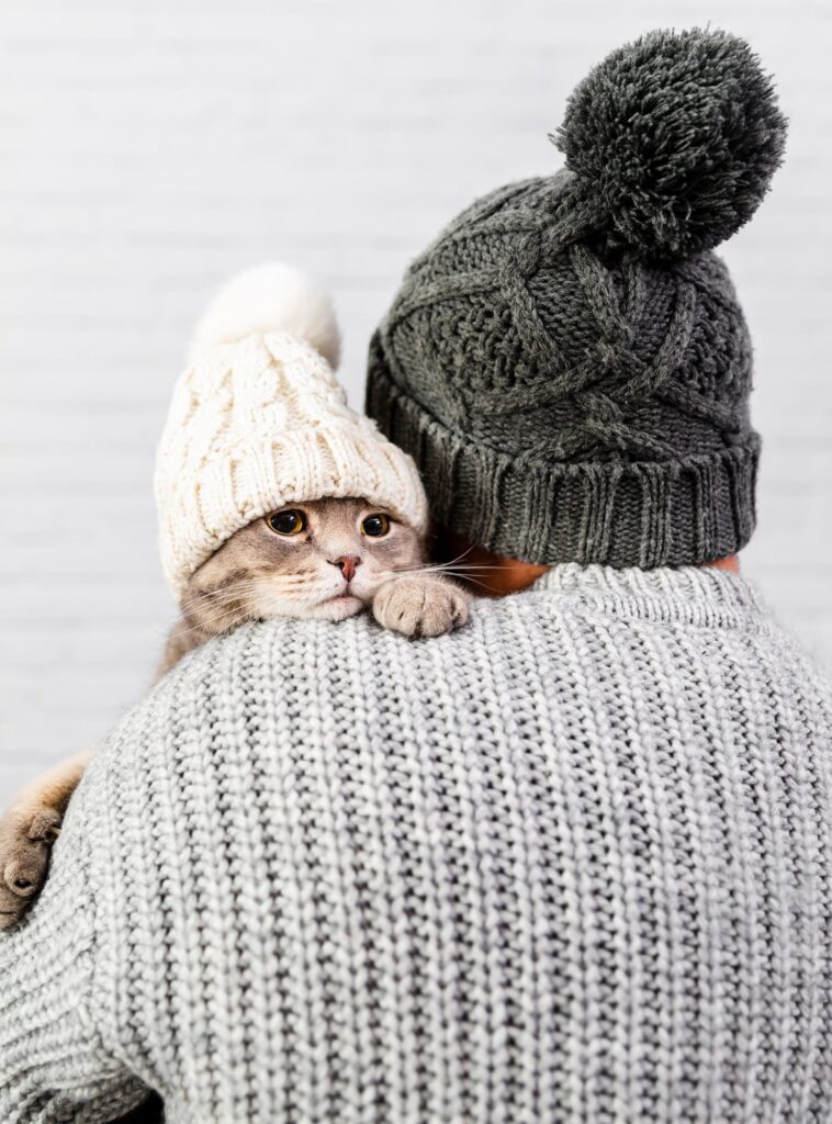 Man in winter attire lovingly holding a cute cat wearing a warm hat, illustrating pet owners' preparation for cold weather conditions to ensure their pets' comfort and safety.